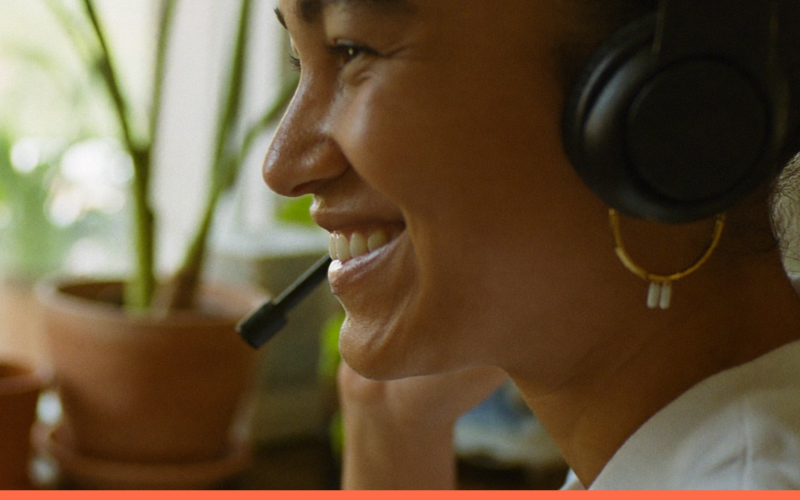 Profissional sorridente com fones de ouvido trabalhando de casa, em mesa de madeira junto à janela com plantas