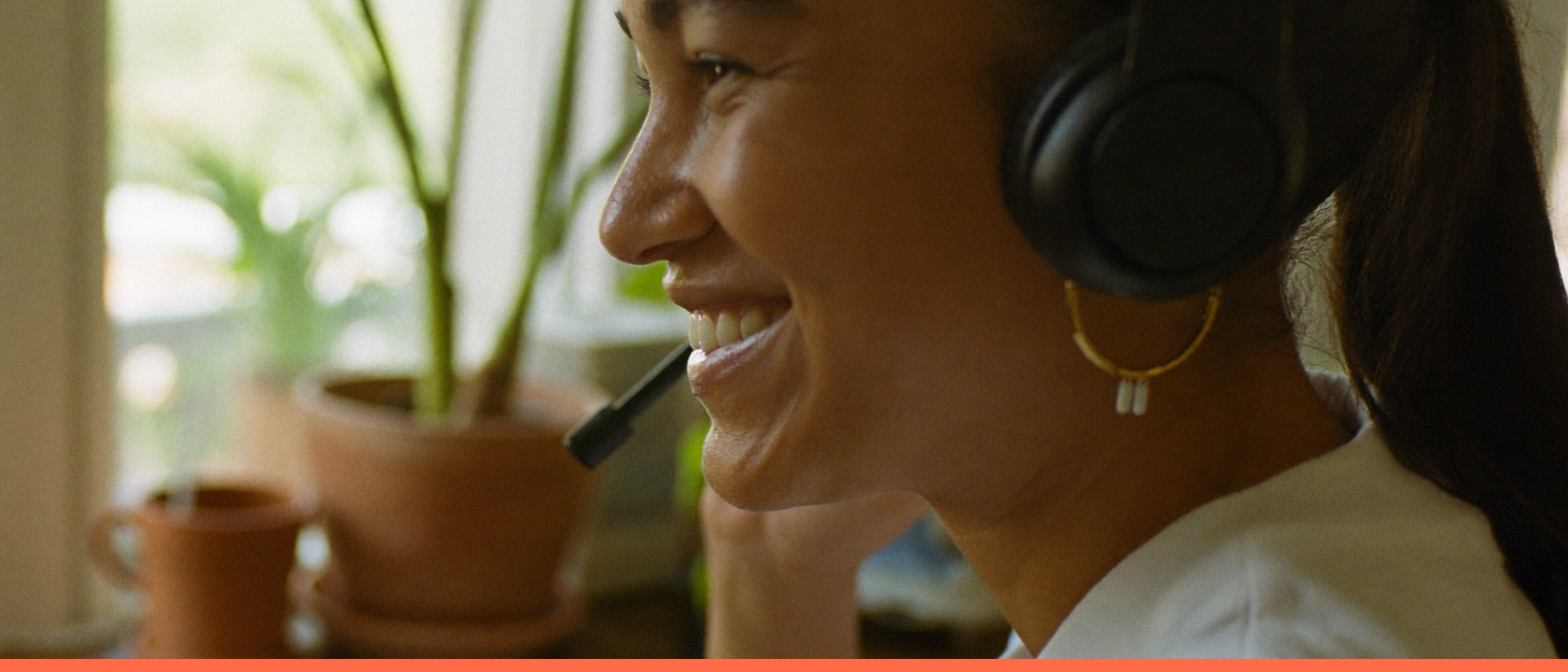 Profissional sorridente com fones de ouvido trabalhando de casa, em mesa de madeira junto à janela com plantas