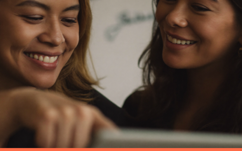 Duas colegas sorrindo enquanto uma mostra a outra o trabalho em um tablet