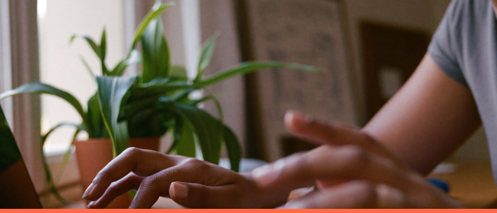Mãos digitando em um notebook sobre mesa clara, com plantas e luz de janela ao fundo